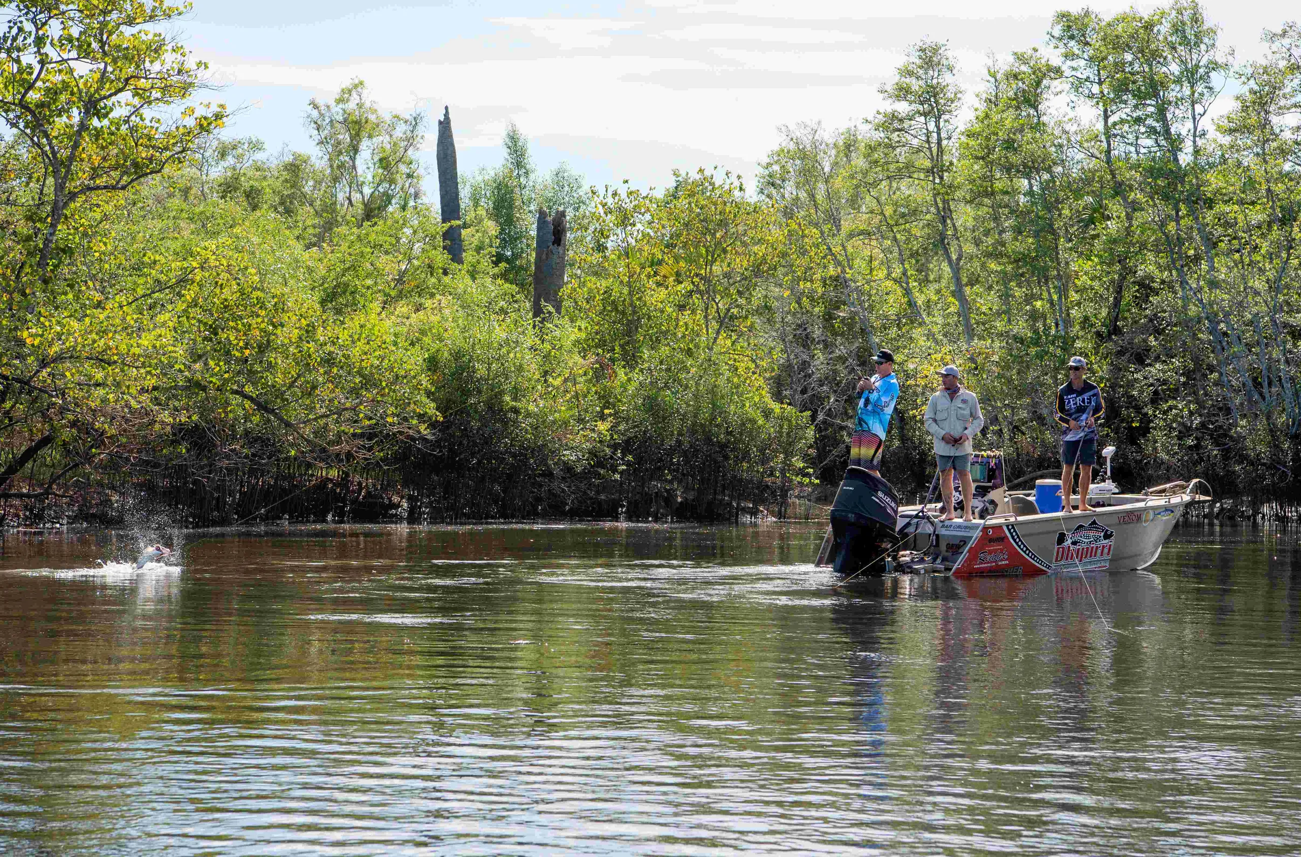 Anglers fish from a boat in a mangrove-lined Arnhem Land creek as a fish breaks the surface, with towering termite mounds visible among the trees.