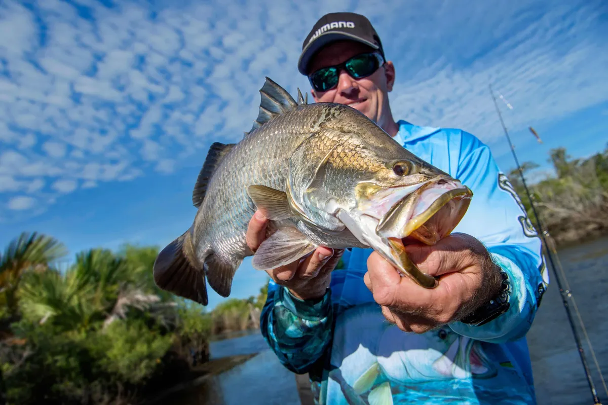 A recreational angler holds a large barramundi toward the camera on a river, with lush tropical vegetation along the banks and a bright blue sky filled with scattered clouds in the background. The fish is prominently displayed in the foreground, showing its silver scales and open mouth, while fishing rods are visible behind the angler.