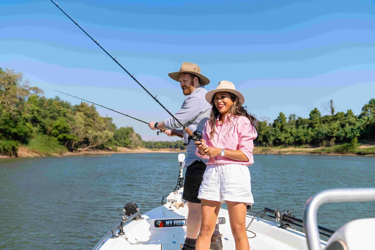 Two people stand on the bow of a fishing boat casting lines into a wide river bordered by dense green bushland. Both are wearing broad-brimmed hats and holding fishing rods, with calm water and a clear blue sky creating a bright outdoor setting. The boat is positioned mid-river, highlighting a recreational fishing experience in a natural tropical landscape.